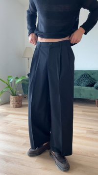 Leveled paisi mustad püksid voldidega, kohandatud kangast, nupukinnitusega ja siledast tekstuurist, koos tumedate jalanõudega.