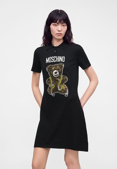 Femme portant une robe polo noire avec un motif d'ours en peluche et le texte "MOSCHINO" sur le devant, debout les mains derrière le dos.