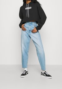 Persona che indossa un hoodie cropped nero di Levi's, jeans chiari a vita alta, calzini bianchi e sneakers Vans nere, in piedi con le mani sui fianchi.