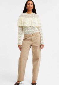 Blonde top med flæset halsudskæring i creme, parret med beige højtaljede vidde bukser. Sorte hæle afslutter outfittet.