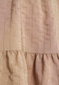 Tissu texturé brun clair avec des fils métalliques verticaux et un détail de volants froncés sur le bord inférieur.