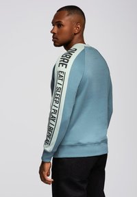 Sudadera azul claro de mangas largas, con un diseño de texto negro vertical en la manga izquierda. La tela tiene una textura suave.