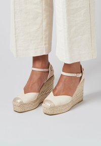 Sandales compensées en espadrille beige avec une semelle en jute texturé, une tige en tissu et un bout doré brillant, dotées d'une bride de cheville.
