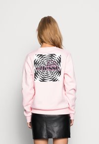 Sudadera rosa de manga larga, con un gráfico de espiral hipnótica en blanco y negro en la parte posterior. Tejido suave, corte relajado.