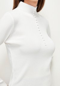 Haut blanc à manches longues avec un col montant, comprenant une rangée de boutons décoratifs blancs sur le devant, fabriqué en tissu doux et texturé.