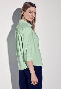 Grön kragebomullsskjorta med uppvikta 3/4 ärmar, sidoslit och bärs med mörkblå byxor, har en mjuk textur och avslappnad passform.