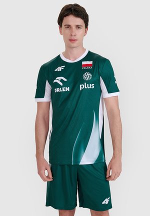 Młody mężczyzna stojący, ubrany w zieloną sportową koszulkę i szorty Polski z logo sponsorów na jednolitym tle.