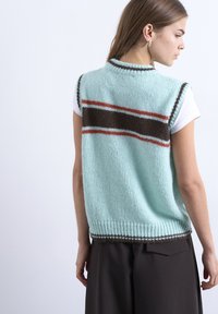 Νεαρή γυναίκα με μακριά καστανά μαλλιά φοράει ανοιχτό μπλε πλεκτό γιλέκο χωρίς μανίκια με καφέ και κόκκινες ρίγες πάνω από λευκό κοντομάνικο πουκάμι σο.