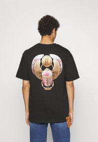 Schwarzes T-Shirt mit einem farbenfrohen Skarabäus-Design auf dem Rücken, mit goldenen Akzenten und Flügelmotiven. Enthält ein orangefarbenes Markenschild an der Seite.