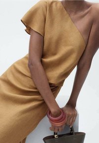 Braunes, asymmetrisches Kleid mit kurzen Ärmeln, aus einem strukturierten Stoff gefertigt. Das Model trägt farbenfrohe Armreifen und hält eine dunkle Handtasche.