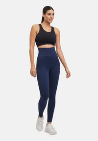 Zwarte sportbeha en hoge marineblauwe legging met een textuurpatroon, gecombineerd met witte sportschoenen. Model is aan het lopen.