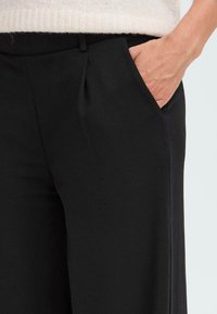 Pantalon noir ajusté avec une texture lisse, doté d'une poche latérale, de légères plis à la taille et d'une coupe droite.