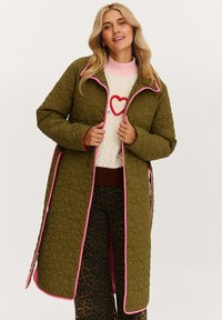Prešívaný dlho zelený kabát s ružovým lemom a zapínaním na cvoky, so štruktúrovanou tkaninou a bočnými rozparkami, nosený cez vzorovaný sveter.