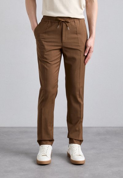 Pantalons marron, coupe droite, avec une taille élastique et un cordon de serrage. Comprend des poches latérales et un tissu lisse et léger. Portés avec des baskets blanches.