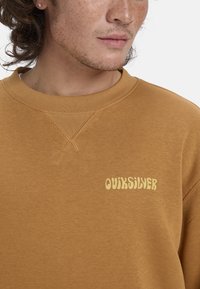 Felpa in cotone marrone con scollatura a coste e logo "QUIKSILVER" ricamato in giallo. Presenta cuciture visibili e una vestibilità comoda.