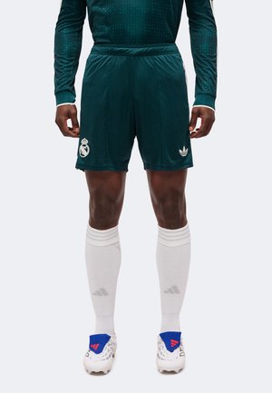 Mężczyzna sportowiec w ciemnozielonych krótkich spodenkach piłkarskich z logo Real Madryt i Adidas, białych skarpetach do kolan oraz białych korkach z niebieskimi i czerwonymi detalami.