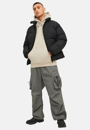 Młody mężczyzna stoi z rękami w kieszeniach, ubrany w beżową bluzę z kapturem, czarną kurtkę puchową, szare spodnie typu cargo oraz czarne sneakersy na białym tle.