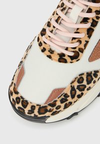 Μύτη αθλητικού παπουτσιού με λευκό ύφασμα, γούνινη επένδυση με σχέδιο λεοπάρ, λεπτομέρειες από ανοιχτό καφέ δέρμα και ροζ κορδόνια σε άσπρη σόλα με μαύρη αντιολισθητική βάση.