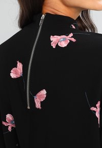 Musta kangasmekko, jossa on vaaleanpunaisia kukkaompeluja. Mekossa on korkea kaulus ja takana näkyvä metallivetoketju.