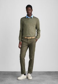 Olive green sweater over a light blue collared shirt, paired with olive trousers and white sneakers. Smooth texture, casual fit.