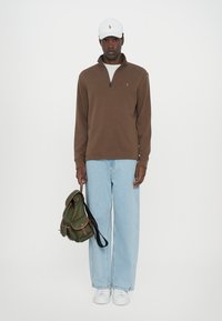 Bruine rits-pullover, lichtblauwe wijde jeans, witte sneakers, groene rugzak met bruine accenten en witte pet met logo.