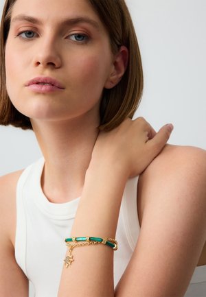 Bracelet en or avec des accents de barres vertes et un charme en forme de soleil. Le modèle porte un haut sans manches blanc, mettant en valeur les bijoux à son poignet.
