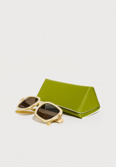 Lunettes de soleil carrées de couleur crème avec des verres foncés à côté d'un étui en cuir triangulaire vert citron sur fond blanc.