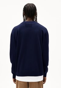 Pull en tricot bleu marine avec une coupe décontractée, des poignets côtelés et un patch texturé dans le dos. Superposé sur une chemise blanche, associé à un pantalon beige.