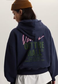 Marineblauwe hoodie met een gestructureerde afbeelding in roze en groen met de tekst "Vans OFF THE WALL." Heeft een losse pasvorm en een verstelbare capuchon.
