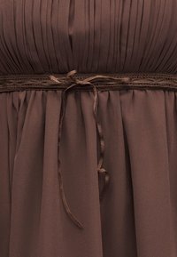 Καφέ ύφασμα για φόρεμα με κάθετες πιέτες πάνω από μια λεπτή πλεγμένη ζώνη δεμένη με φιόγκο στη μέση, με μαλακές πτυχώσεις κάτω από αυτήν.