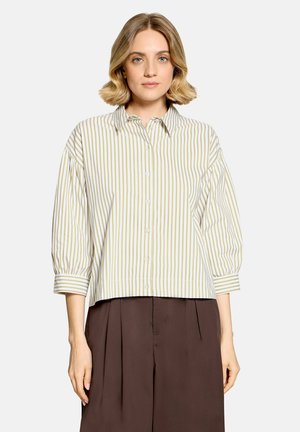 Woman with short blonde hair wearing a beige and white vertical striped button-up shirt and loose brown pants standing against a plain background.