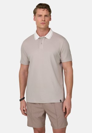 Mand iført beige kortærmet poloshirt med hvid krave og mønstrede beige shorts står mod hvid baggrund.