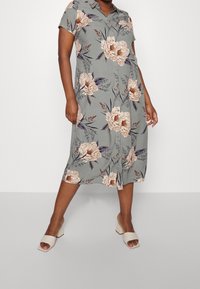 Blumenkleid aus grauem Stoff mit kurzen Ärmeln, verziert mit großen beigen und lila Blumen und einer durchgehenden Knopfleiste. Getragen mit beigen High-Heels.