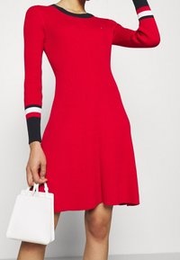 Punainen ribattu neulemekko pitkähihaisilla, kontrastivärisin musta-valkoisin raidoin varustetut hihansuut, liehuva hameosa ja pyöreä kaula-aukko. Valkoinen käsilaukku lisävarusteena.