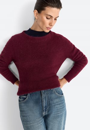 Bordeauxfarbener Rundhalsausschnitt-Pullover mit einer weichen, strukturierten Oberfläche, kombiniert mit hellblauen Jeans, die einen lockeren Schnitt und gerippte Bündchen haben.