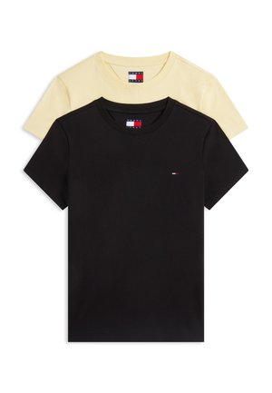Jeune femme aux cheveux bouclés portant un t-shirt jaune à manches courtes, un foulard à motifs autour du cou, un jean bleu à jambes larges avec une ceinture marron et des chaussures foncées.