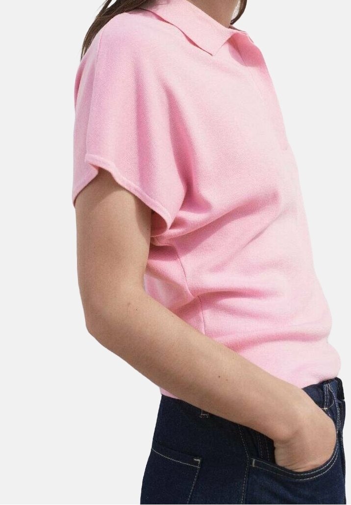 Personne portant une chemise rose pâle à manches courtes avec col, une main dans la poche d’un jean bleu foncé, vue de profil.