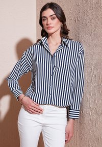 Navy and white striped button-up shirt with a classic collar and long sleeves, paired with white fitted pants. Smooth fabric finish.