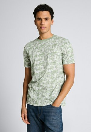 Jonge man draagt een lichtgroen shirt met korte mouwen en een wit bladmotief, staat met één hand in de zak van zijn blauwe spijkerbroek.