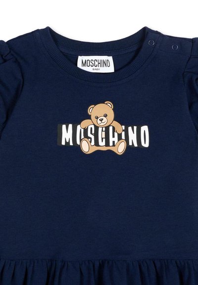 Robe bébé bleu marine avec manches à volants, étiquette Moschino, et graphique d'ours en peluche assis au-dessus du texte blanc « MOSCHINO » sur le devant.