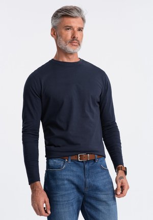 Camicia a maniche lunghe blu abbinata a jeans blu. La camicia presenta un collo rotondo e un design attillato, con cintura e orologio in pelle marrone visibili.
