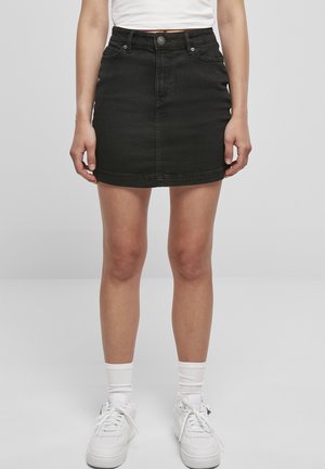 Kvinde iført en sort denim mininederdel, hvide sokker og hvide sneakers, stående med armene afslappet langs siden mod en ensfarvet baggrund.