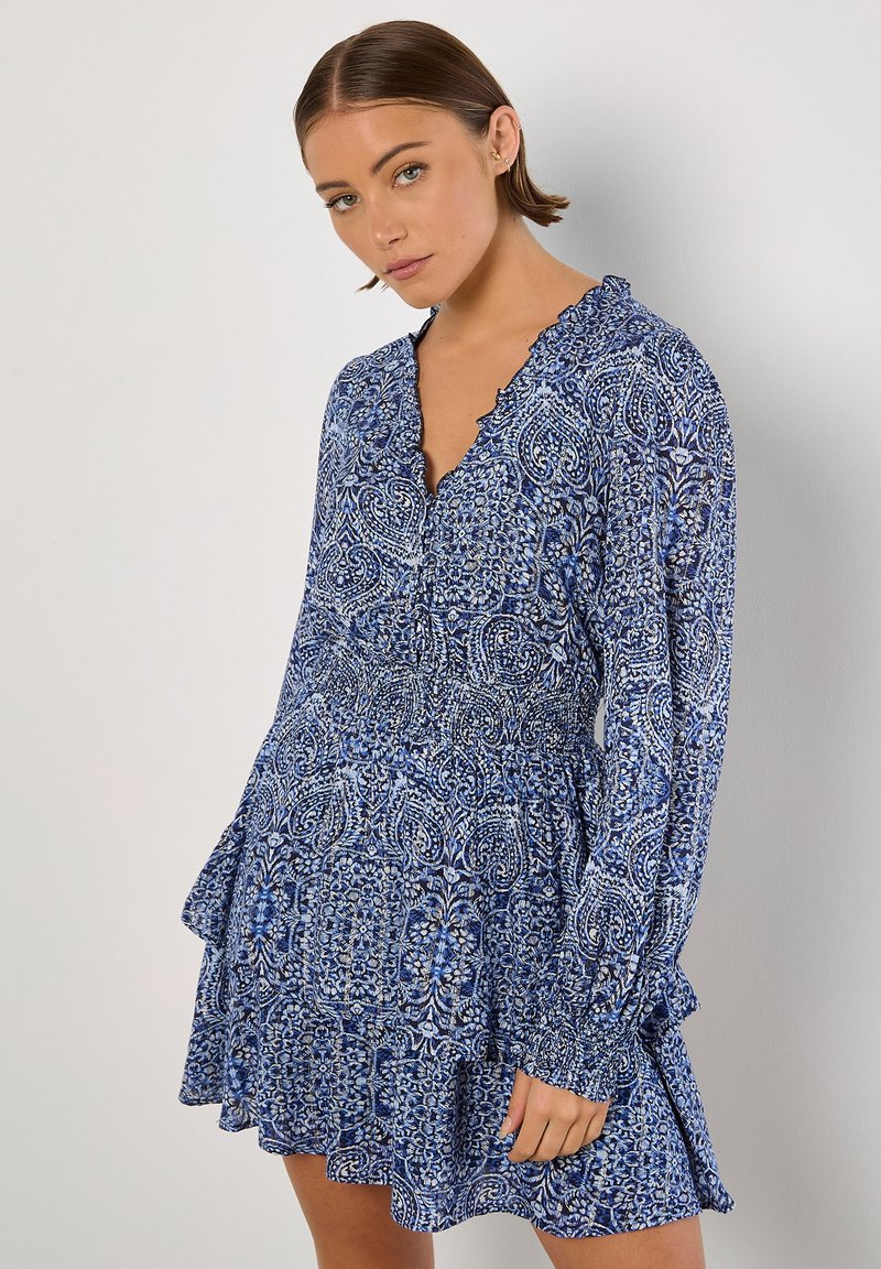 Blau gemustertes Kleid mit langen Ärmeln, rüschenbesetztem Ausschnitt und gestuftem Rock. Der Stoff hat eine glatte Textur mit kunstvollen Designs in verschiedenen Blautönen.