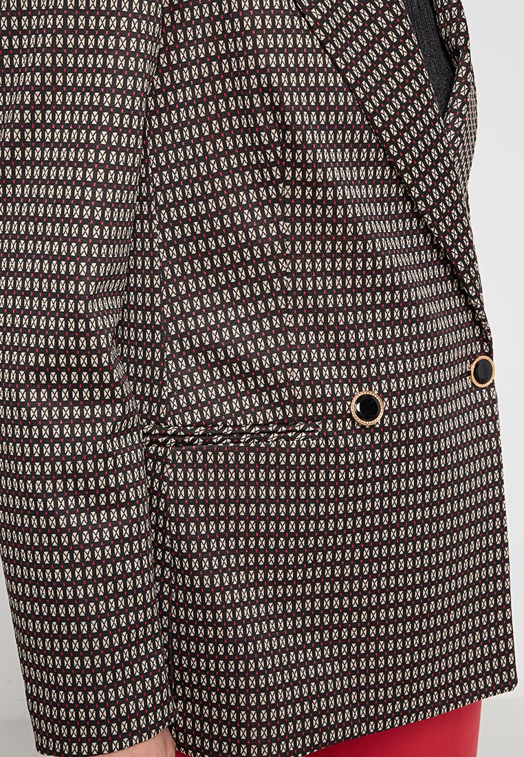Blazer mit geometrischem quadratischem Muster in Schwarz, Beige und Rot, ausgestattet mit schwarzen Knöpfen mit goldenen Kanten, getragen über roten Hosen.