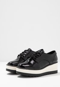 Chaussures à plateforme en cuir noir brillant avec un bout rond, une semelle intermédiaire blanche et une semelle en caoutchouc noir texturée. Elles présentent un design à lacets et des accents de couture.