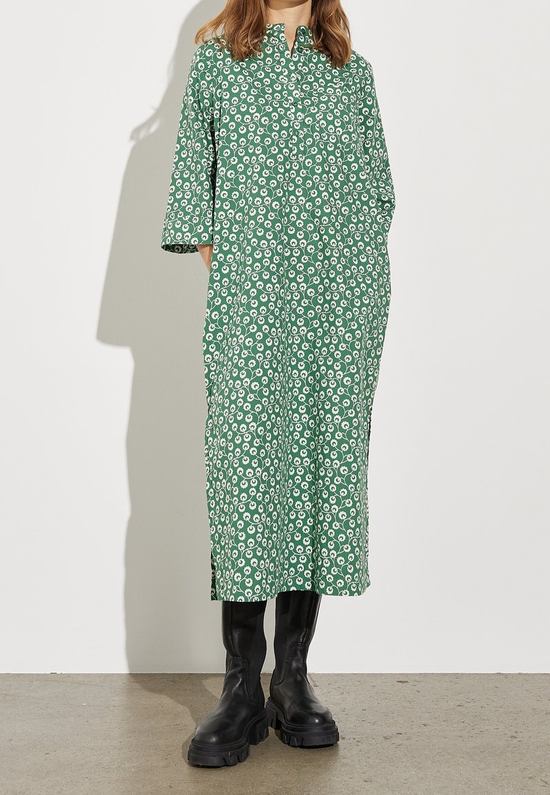 Femme portant une robe verte à motif floral blanc et des bottes noires épaisses, debout devant un fond clair uni.