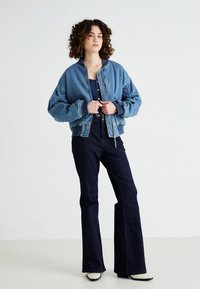 Denim bomber jakna, svijetlo plava, s spuštenim ramenima i elastičnim manžetama; uparena s tamnim flared trapericama i bež čizmama do članaka.