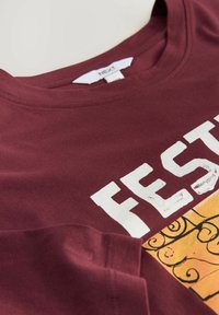 Bordo kokvilnas T-krekls ar apaļu kakla izgriezumu, uz tā atrodas balti teksti "FEST" un dzeltena grafika ar izliektiem melniem rakstiem.