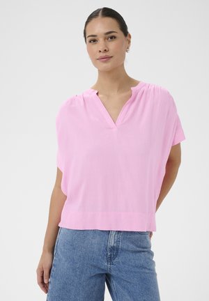 Žena s tmavými vlasmi zviazanými dozadu, nosiaca zlaté kruhové náušnice, voľnú svetloružovú blúzku s výstrihom do V a modré džínsy, stojaca pred jednoduchým pozadím.