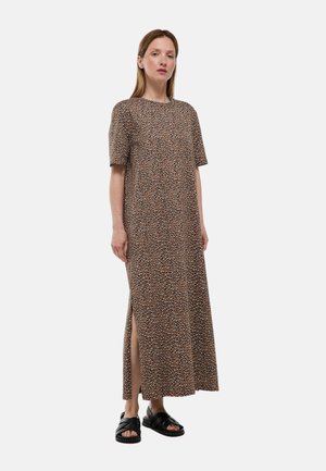 Femme debout portant une robe marron à manches courtes, longueur cheville, avec un petit imprimé et une fente latérale, associée à des sandales noires à bout ouvert.
